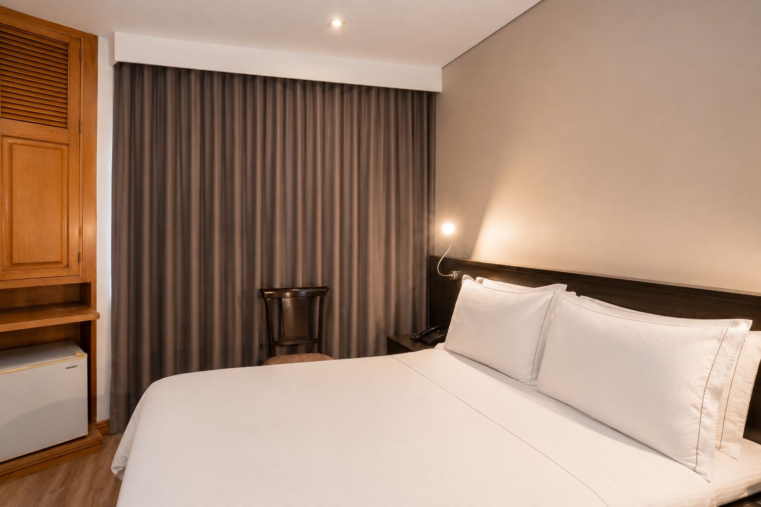 A hotel room with a bed, a wooden cabinet, a chair, and a curtain.