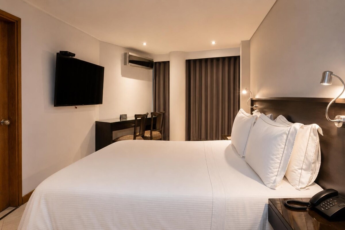 Habitación de hotel con una cama grande, un escritorio con silla, televisor, equipo de aire acondicionado, teléfono y lámpara.