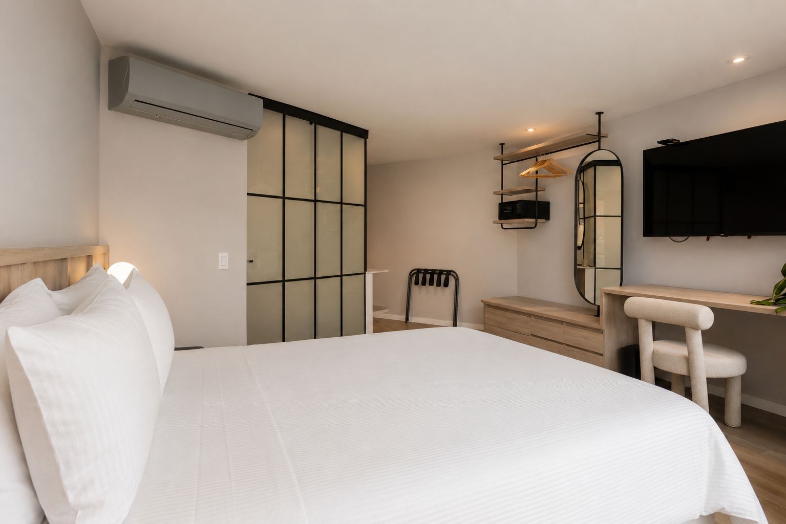 A hotel room with a bed, a desk, a chair, a mirror, and an air conditioning unit.