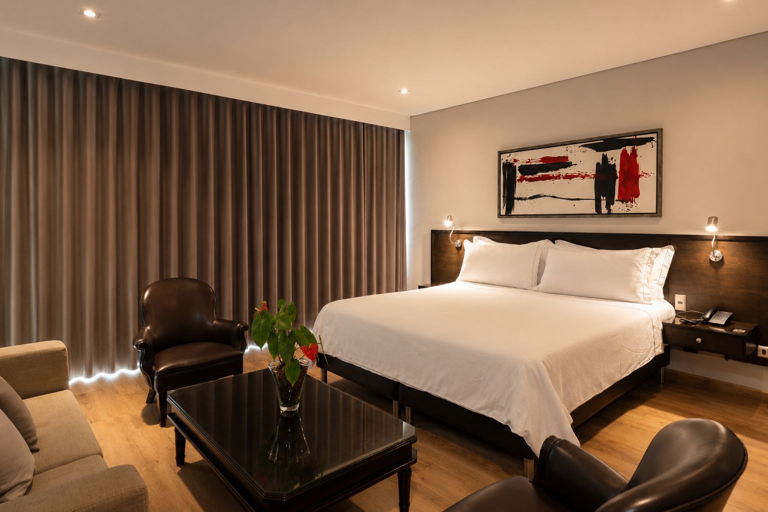 Habitación de hotel con una cama grande, un escritorio, una silla, una mesita de noche y una lámpara de pared.