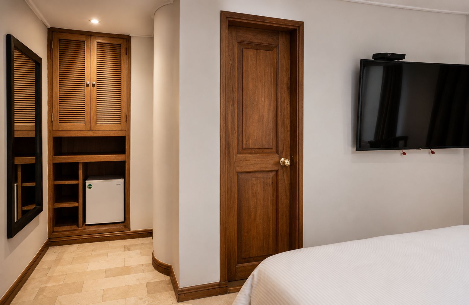 A bedroom with a bed, a wooden wardrobe, a television mounted on the wall, and a white door.