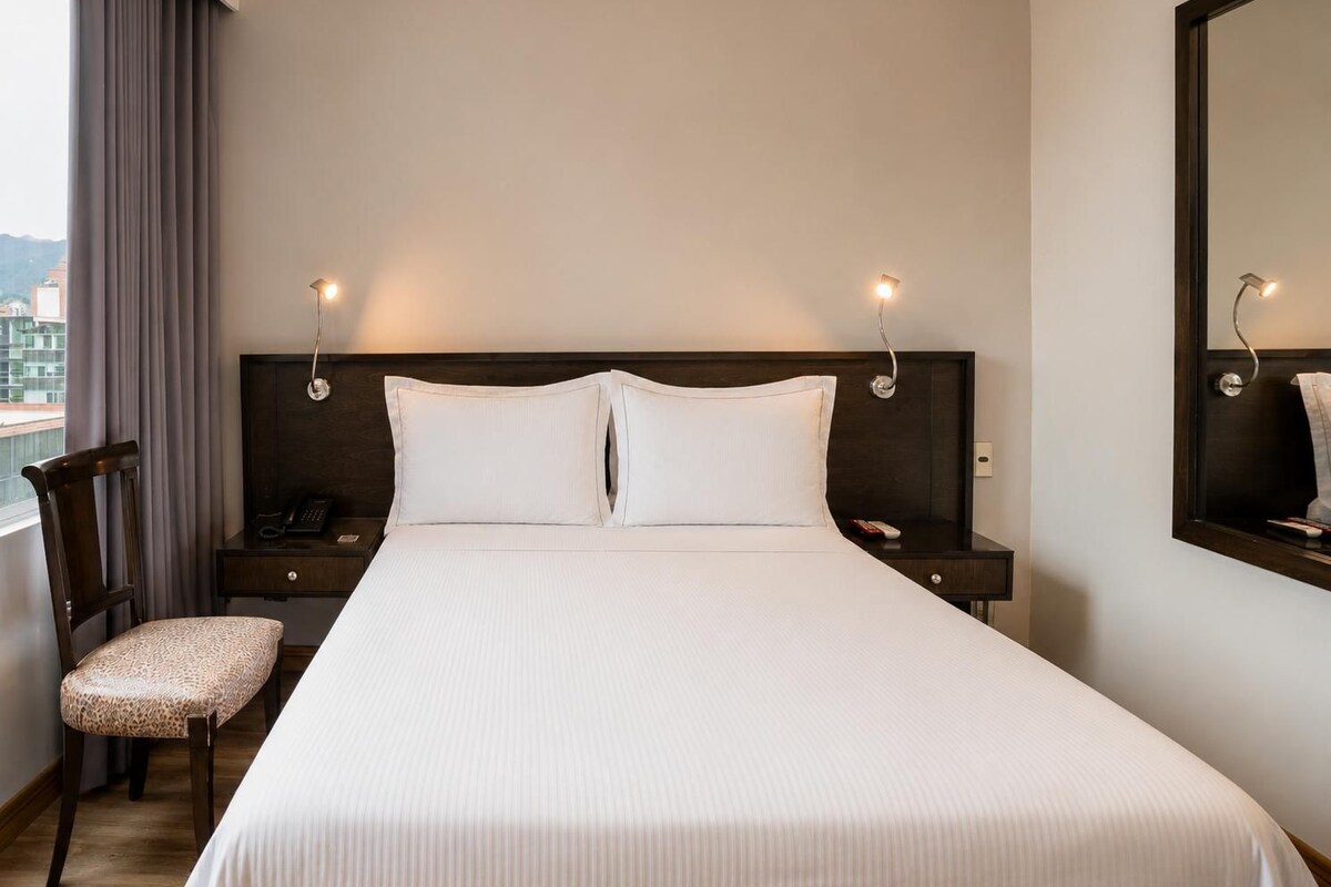 Habitación de hotel con una cama grande, una silla, una mesita de noche con teléfono y un espejo.