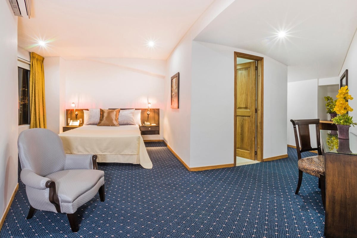 Habitación de hotel con cama, lámparas de noche, una silla, un escritorio y una puerta que da a otra habitación.
