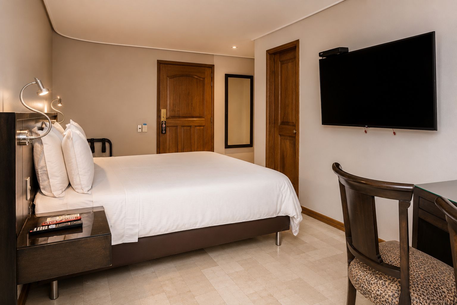 A hotel room with a bed, bedside table, lamp, TV, and a chair.