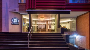 Entrada de hotel con un diseño moderno, que presenta una escalera que conduce al vestíbulo, un candelabro y un letrero con el nombre del hotel.