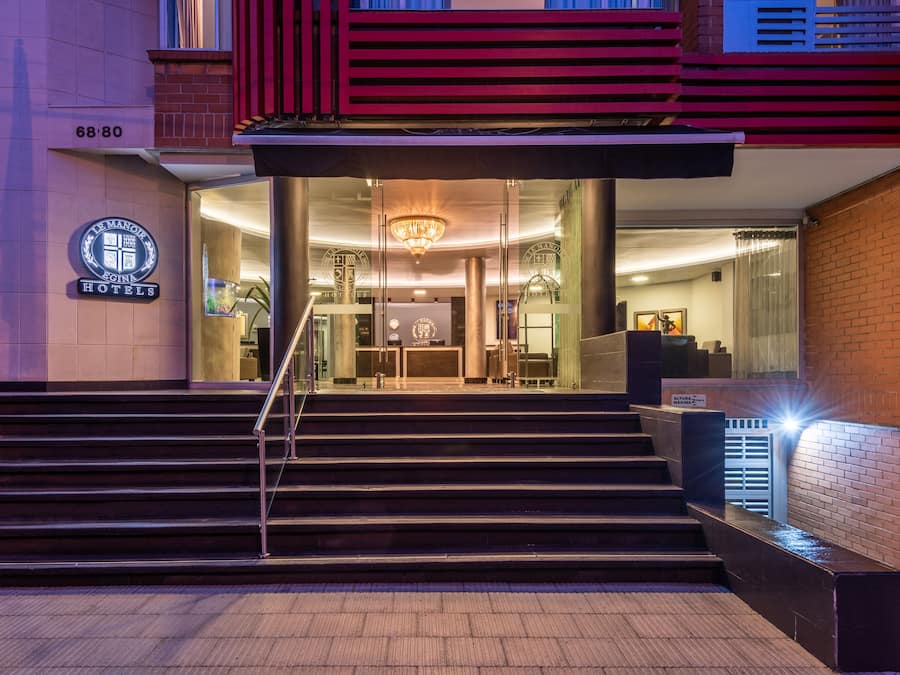 Entrada de hotel con un diseño moderno, que presenta una escalera que conduce al vestíbulo, un candelabro y un letrero con el nombre del hotel.