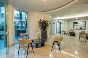 Un vestíbulo de hotel con entrada de vidrio, una zona de estar, una planta en maceta grande y una obra de arte colgada en la pared.