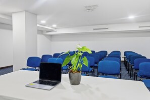Un salón de conferencias con una laptop sobre una mesa, una planta en maceta y filas de sillas azules.