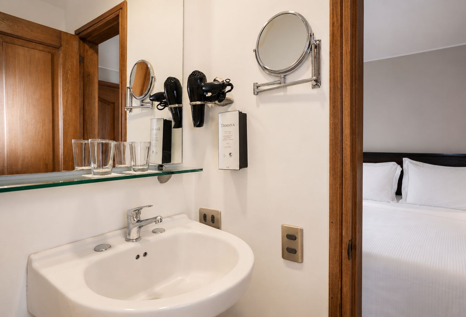 A hotel bathroom with a sink, mirror, hairdryer, and a bed with white bedding.