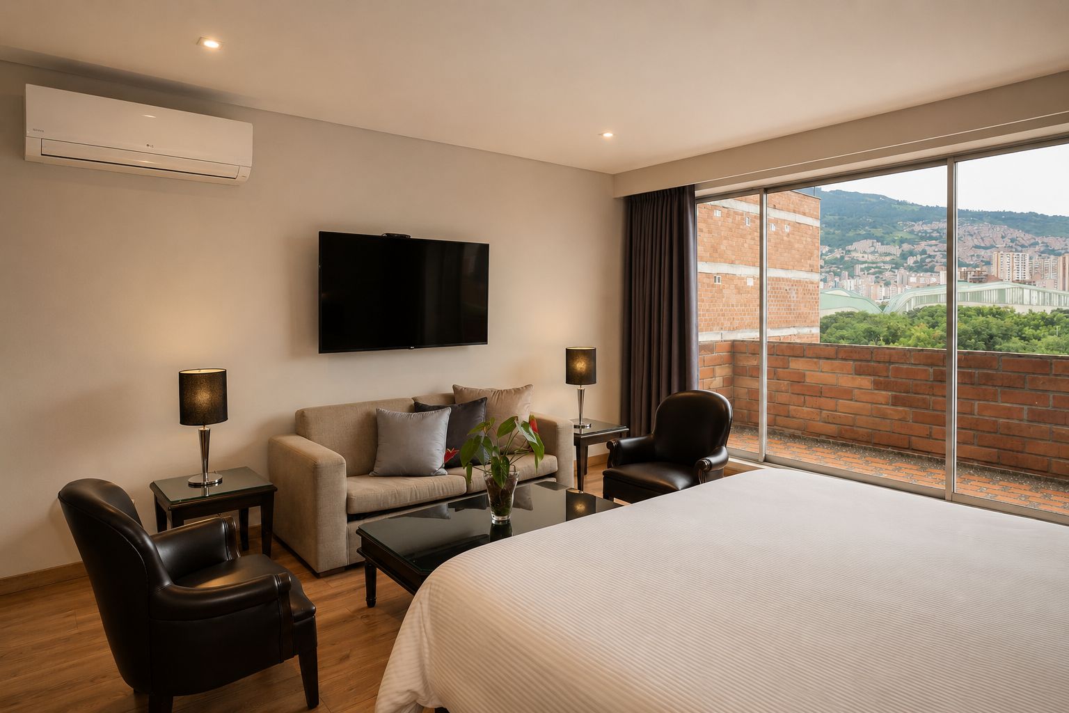 Une chambre d’hôtel comprenant un lit, un canapé, deux fauteuils, une table basse, une télévision et une grande fenêtre donnant sur la ville.