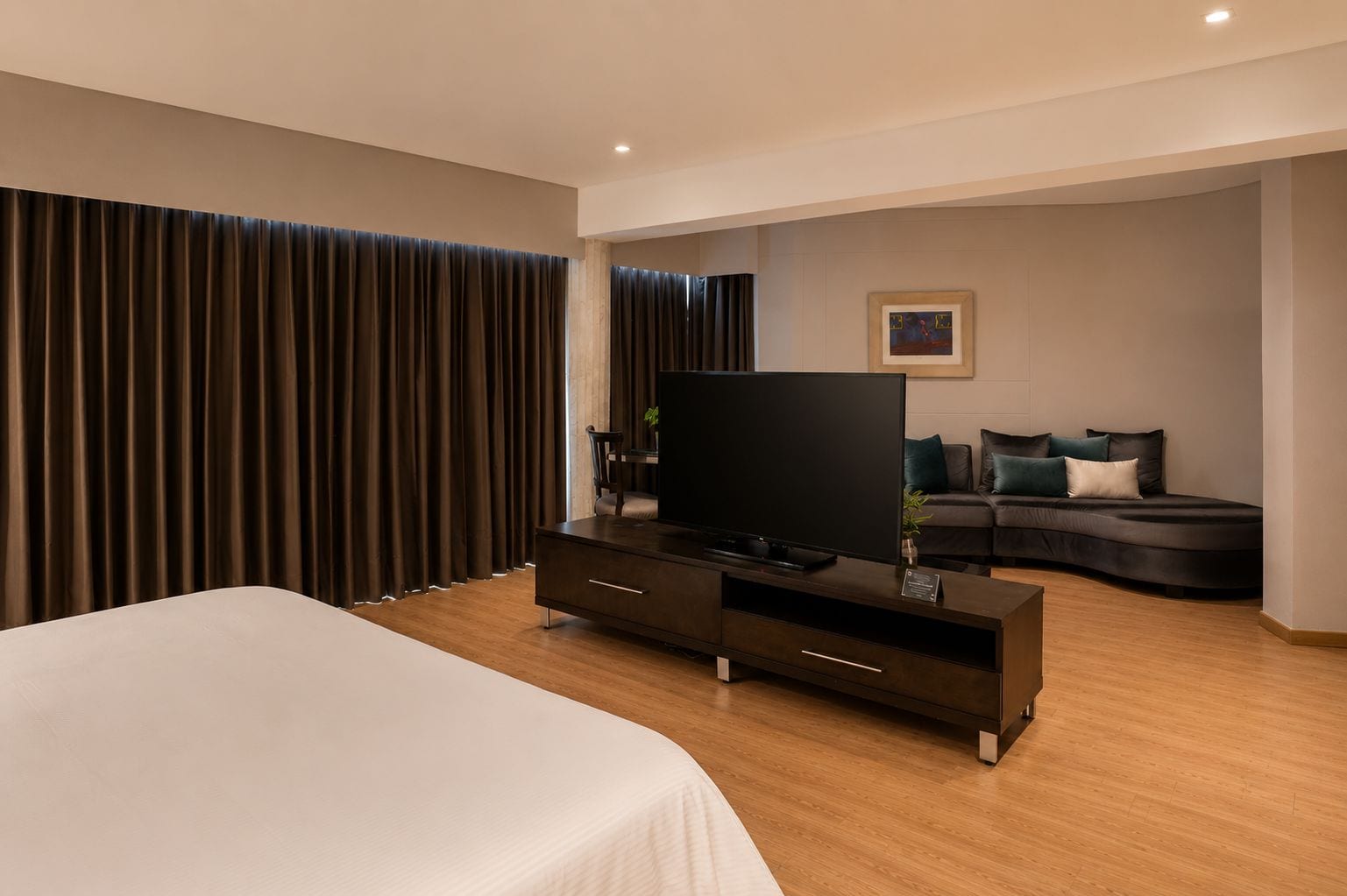 A modern hotel room with a large bed, a flat-screen TV, a dark-colored sofa, and a large window with curtains.