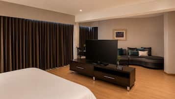 A modern hotel room with a large bed, a flat-screen TV, a dark-colored sofa, and a large window with curtains.