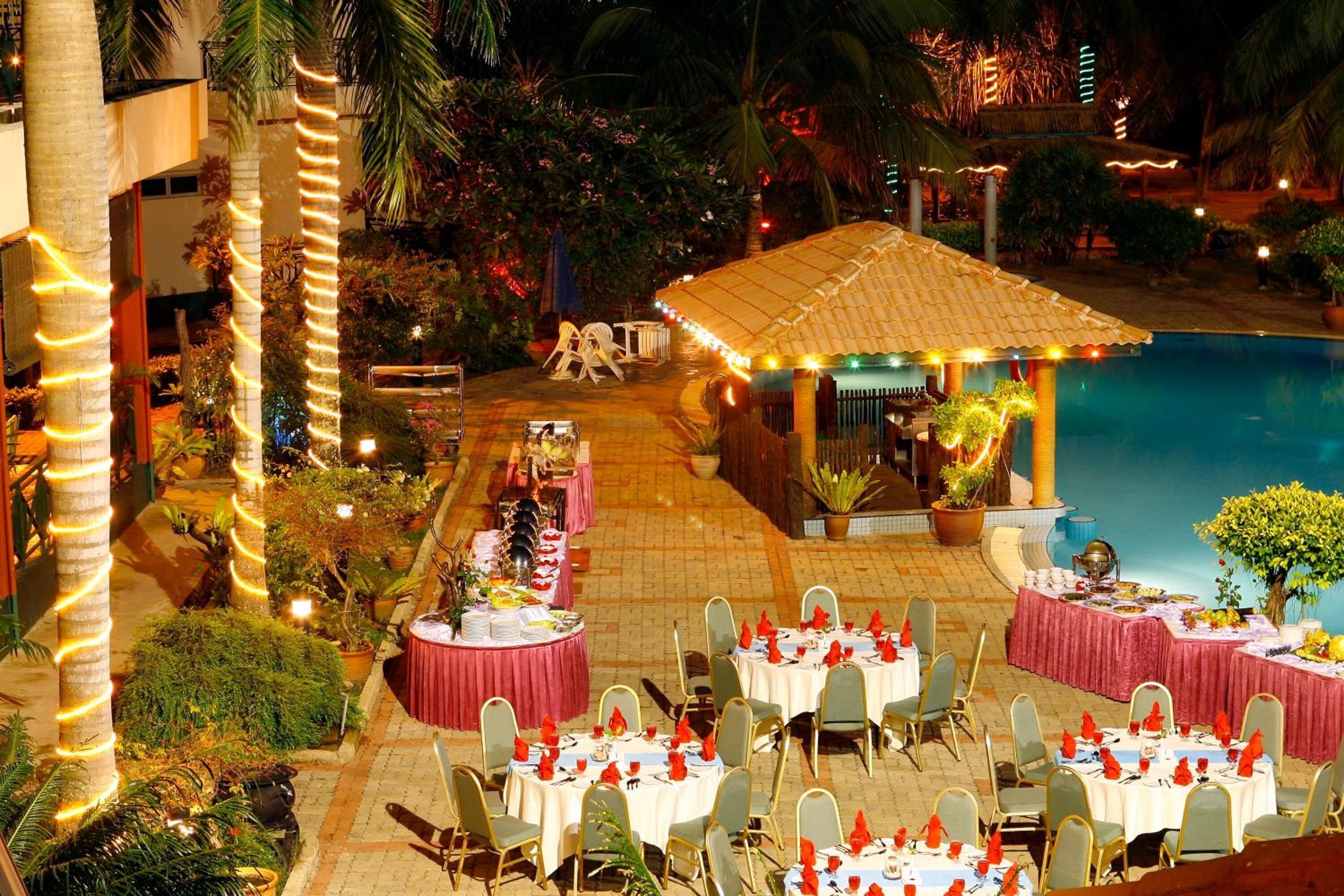 An outdoor dining area with tables set for a meal, surrounded by palm trees and a pool.
