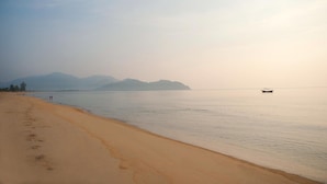 A beach with a boat in the distance, calm water, and mountains in the background. - The Qamar Paka (Dungun)