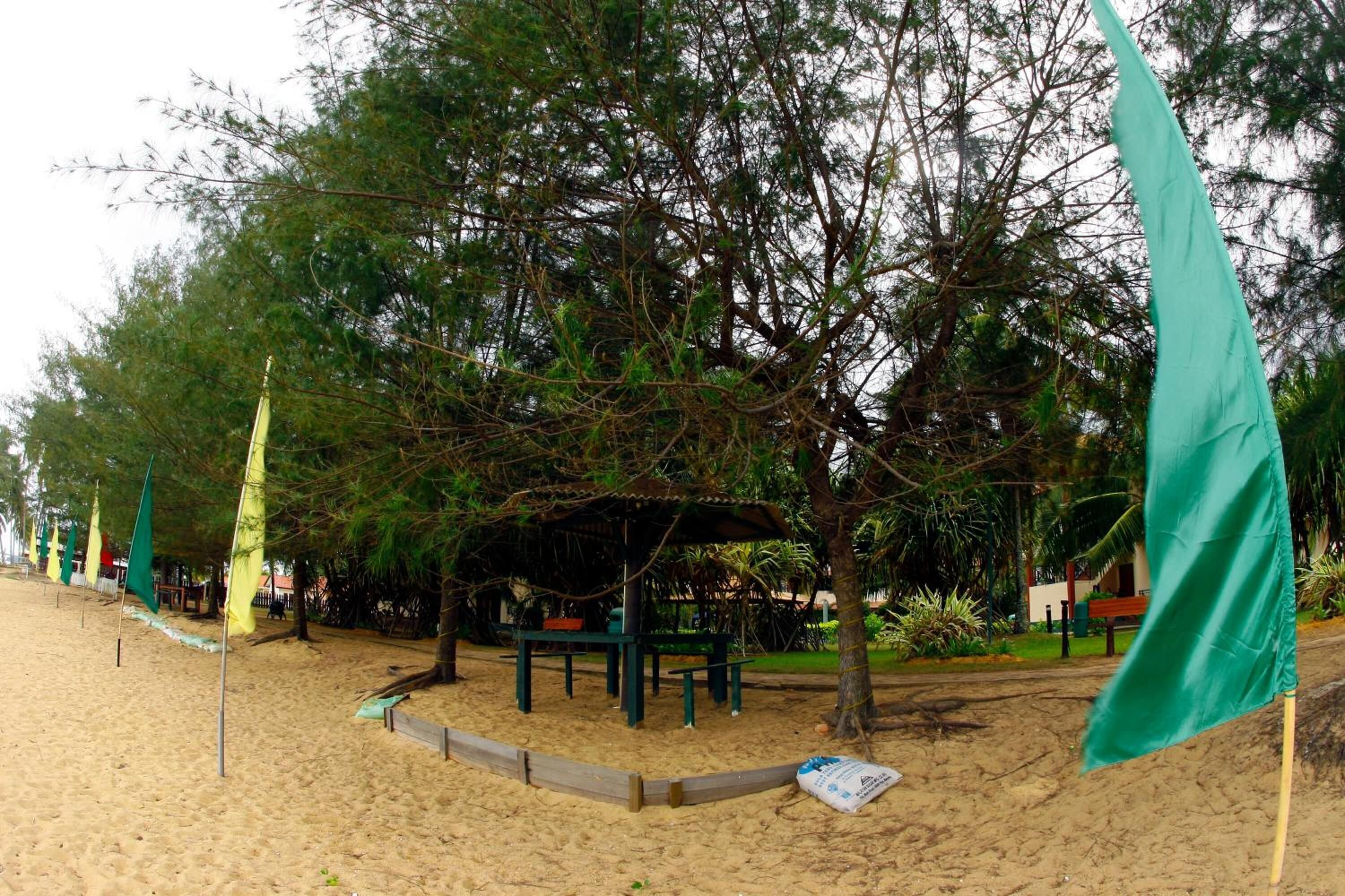 A beach area with a covered seating area, surrounded by trees and flags.