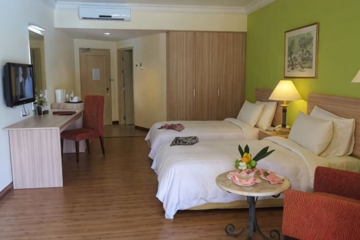 A hotel room with two beds, a desk, a chair, a small table with flowers, and a lamp.