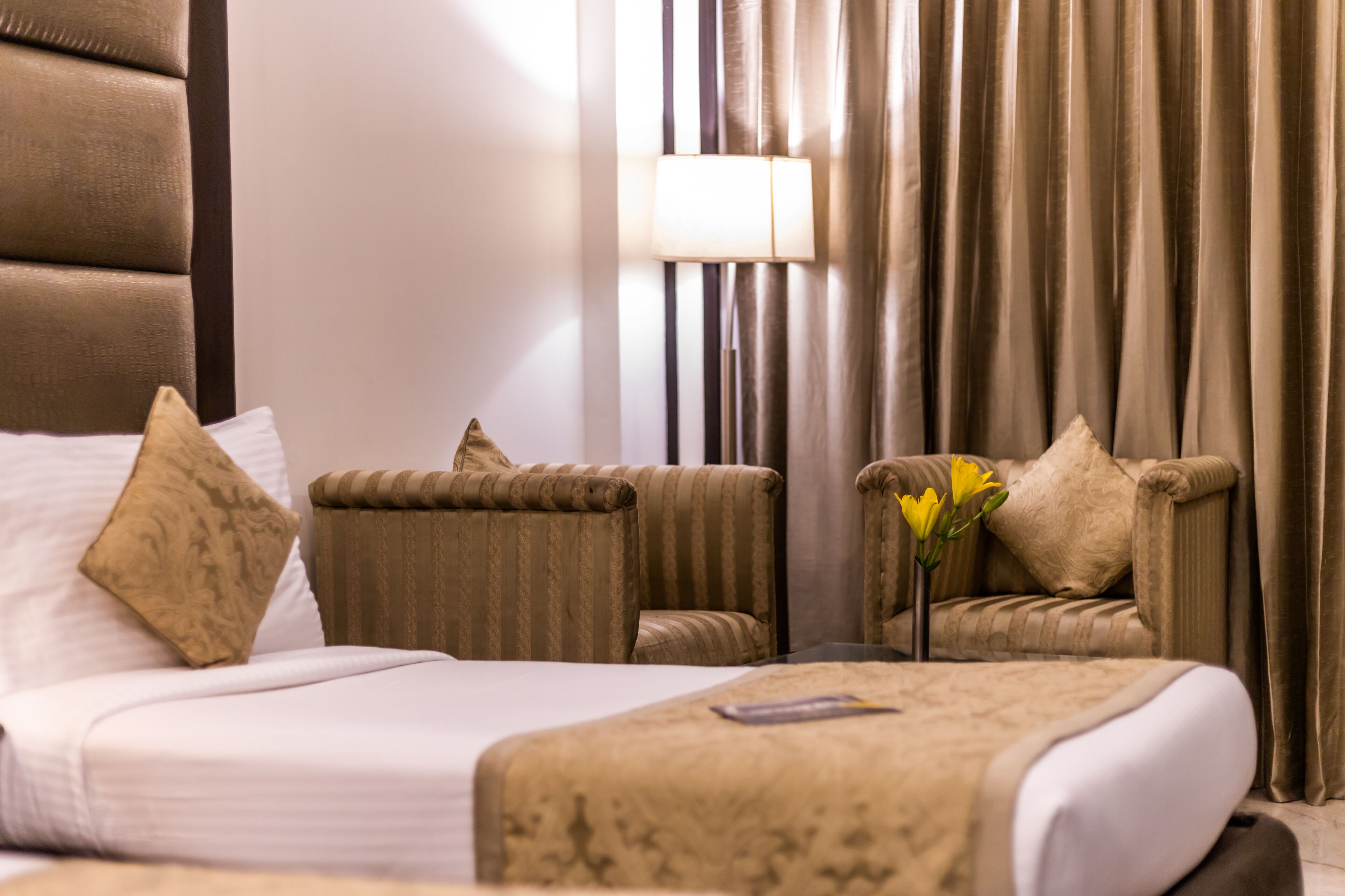 Una habitación de hotel con una cama, dos sillones, una mesita de noche con lámpara y un jarrón con flores.