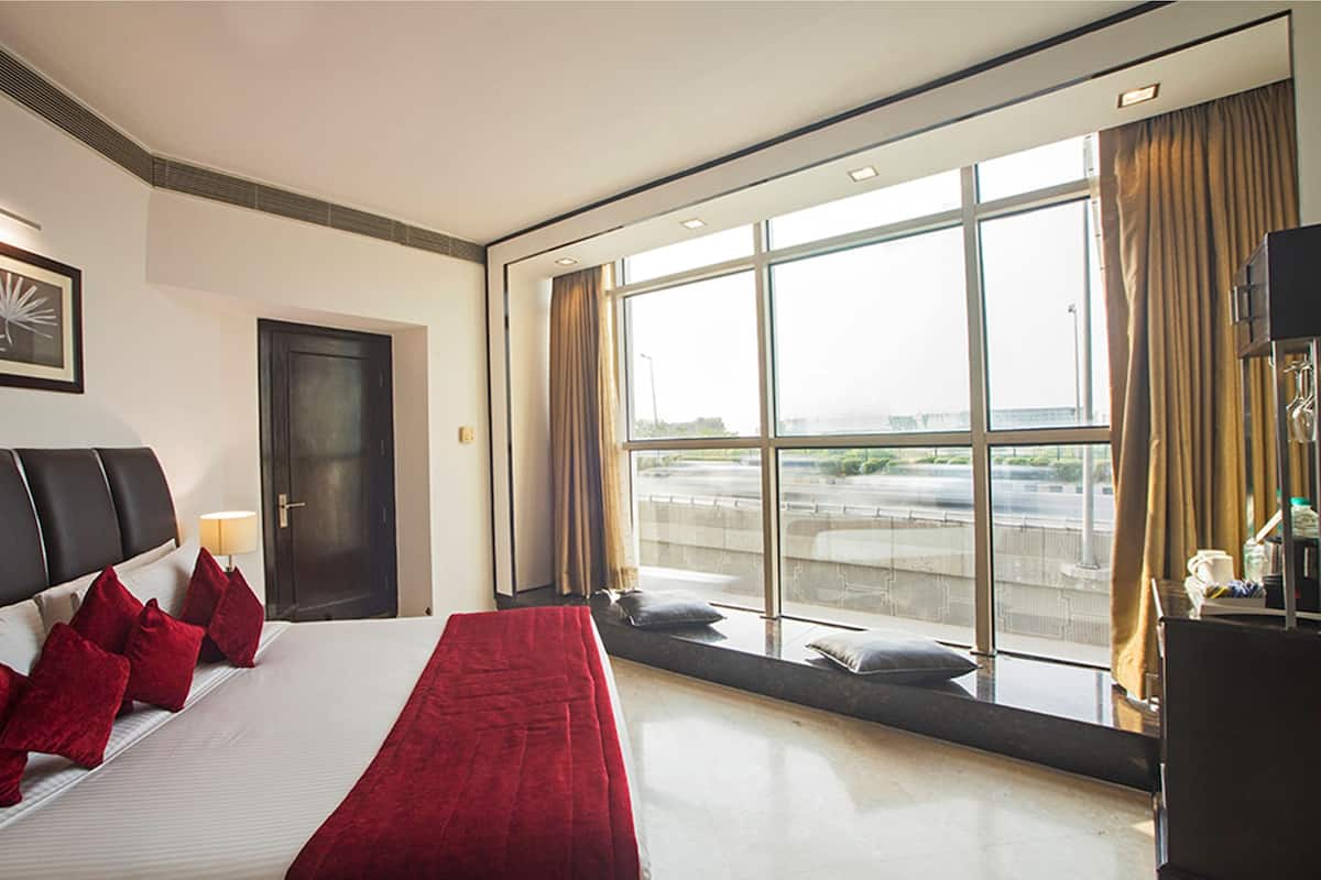 A hotel room with a large bed, a window with a view of the city, a desk with a computer, and a door.