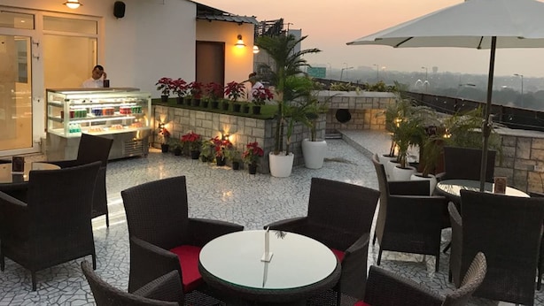 A rooftop terrace with wicker furniture, a glass display case with food items, potted plants, and a view of the cityscape. - Hotel Shanti Palace (New Delhi)