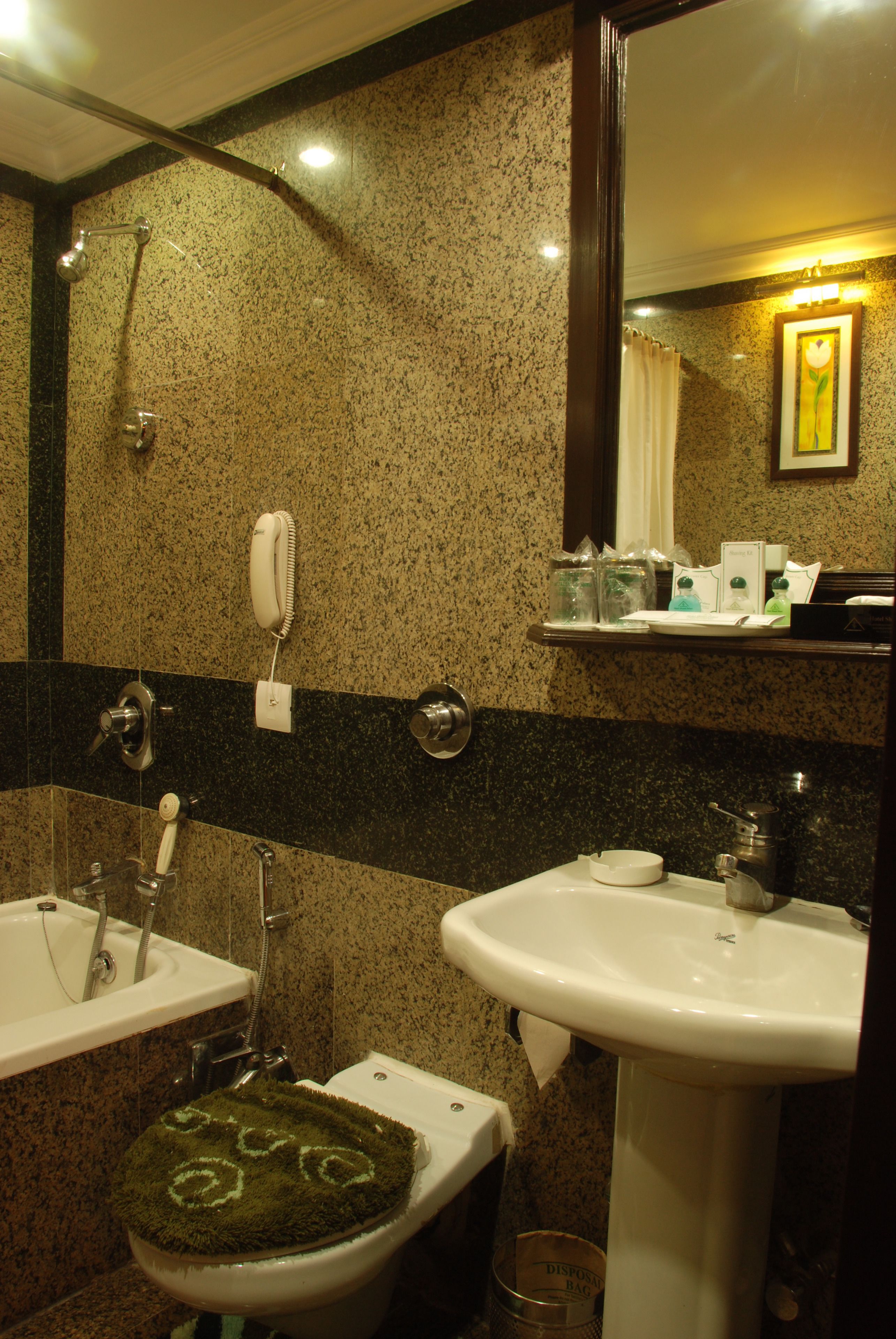 A bathroom with a sink, toilet, and shower. There is a phone on the wall and a trash can.