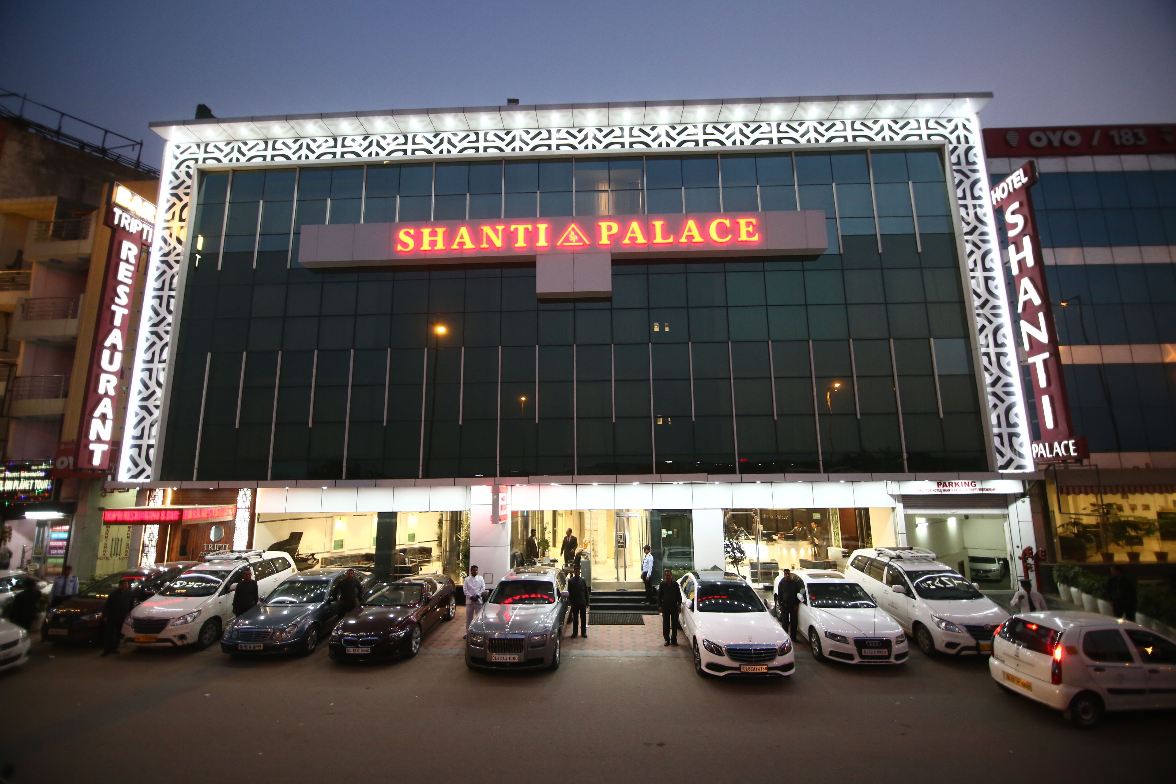 The exterior of Shanti Palace hotel with a modern facade, a prominent entrance, and a parking area with several cars.