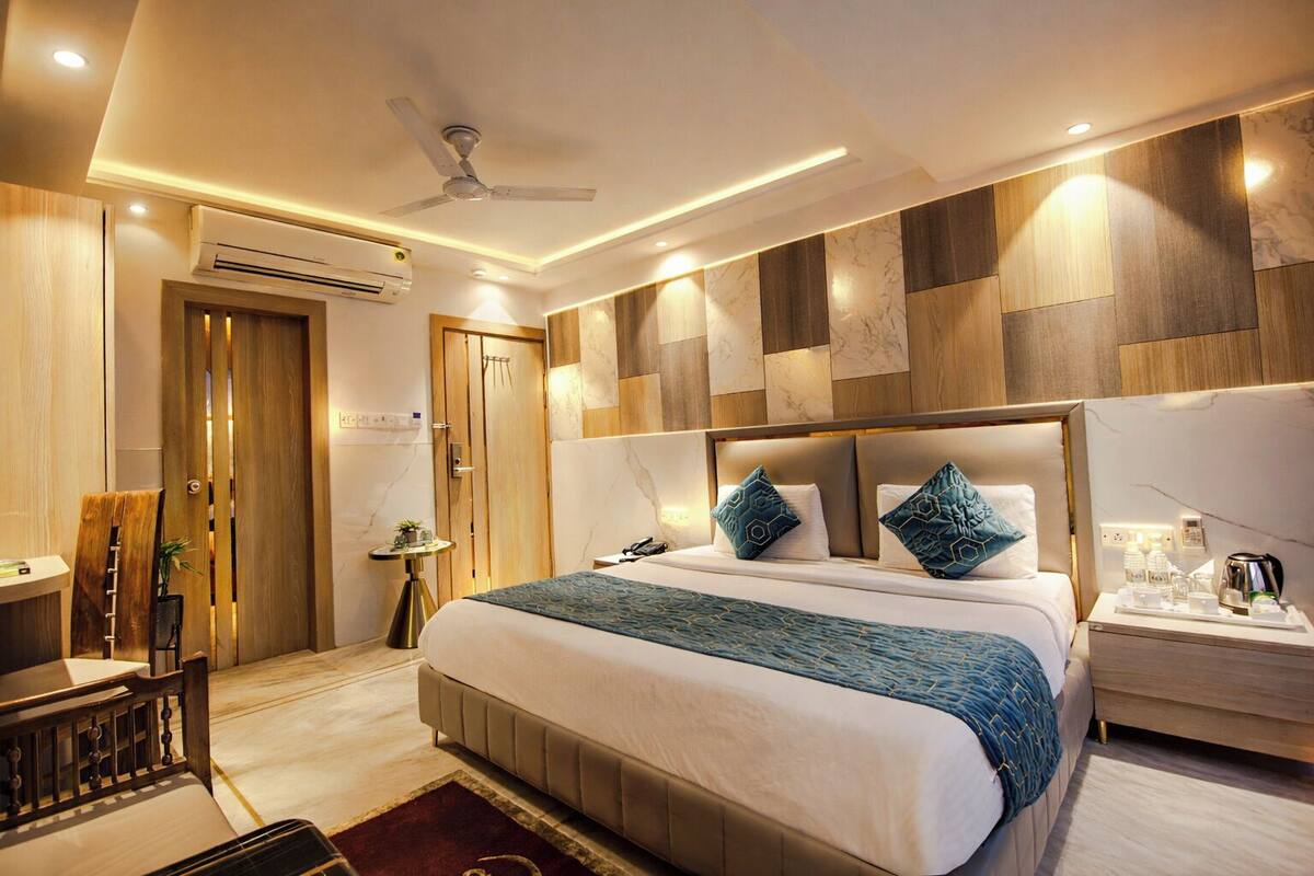 A modern hotel room with a large bed, a bedside table with a lamp, a desk with a chair, and an air conditioning unit.