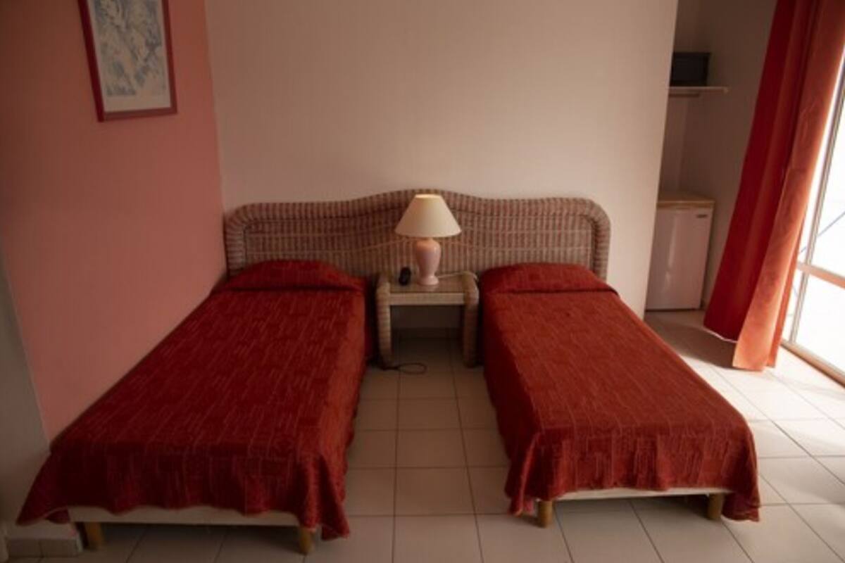 A room with two single beds, a bedside table with a lamp, a small cabinet, and a window with curtains.