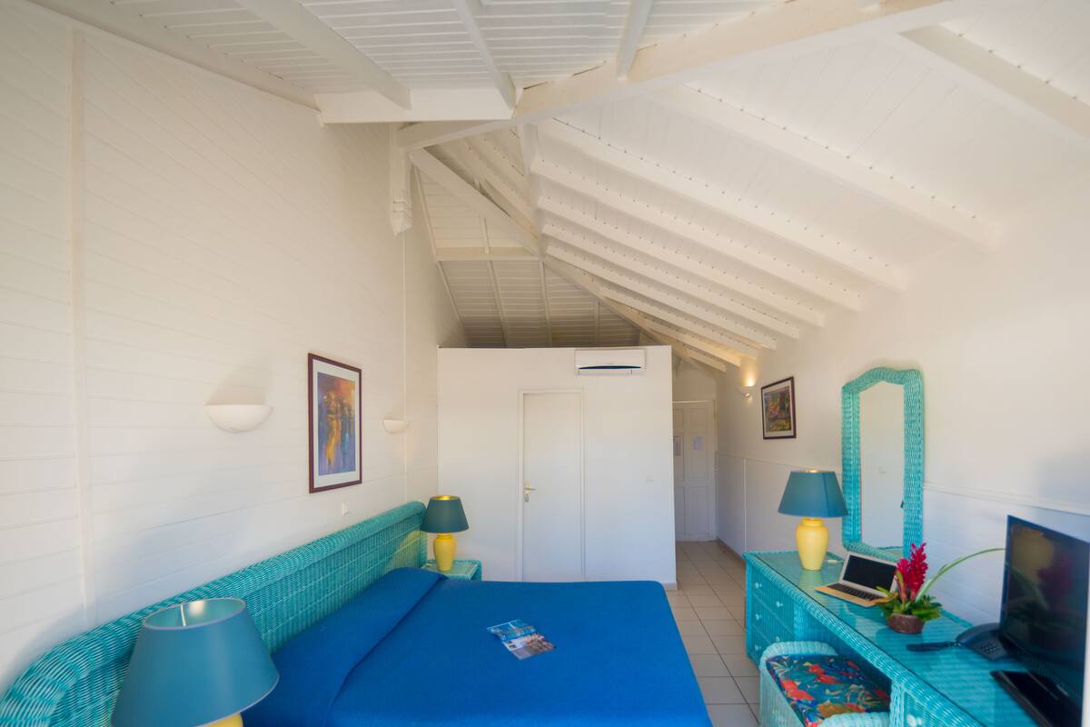 A bedroom with a bed, two bedside tables with lamps, a desk with a computer, a television, and a door leading to another room.