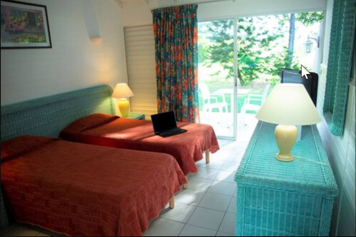 A room with two beds, a laptop on one bed, a lamp on a side table, and a view of greenery outside.