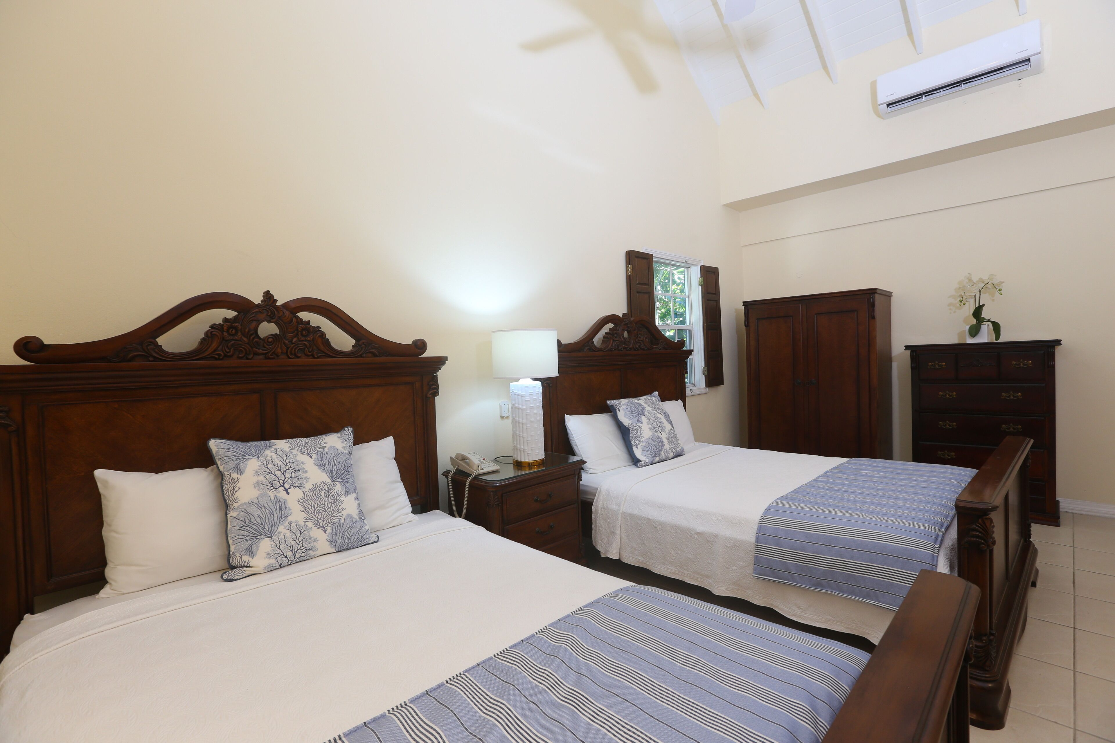 A hotel room with two beds, wooden headboards, a nightstand with a lamp, a dresser, and an air conditioning unit.