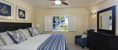 Un dormitorio con una cama, ventilador de techo, ventana con contraventanas, cómoda y espejo.