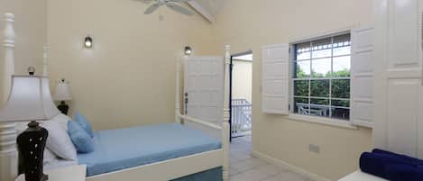 A bedroom with a bed, bedside table, lamp, ceiling fan, and window with shutters.