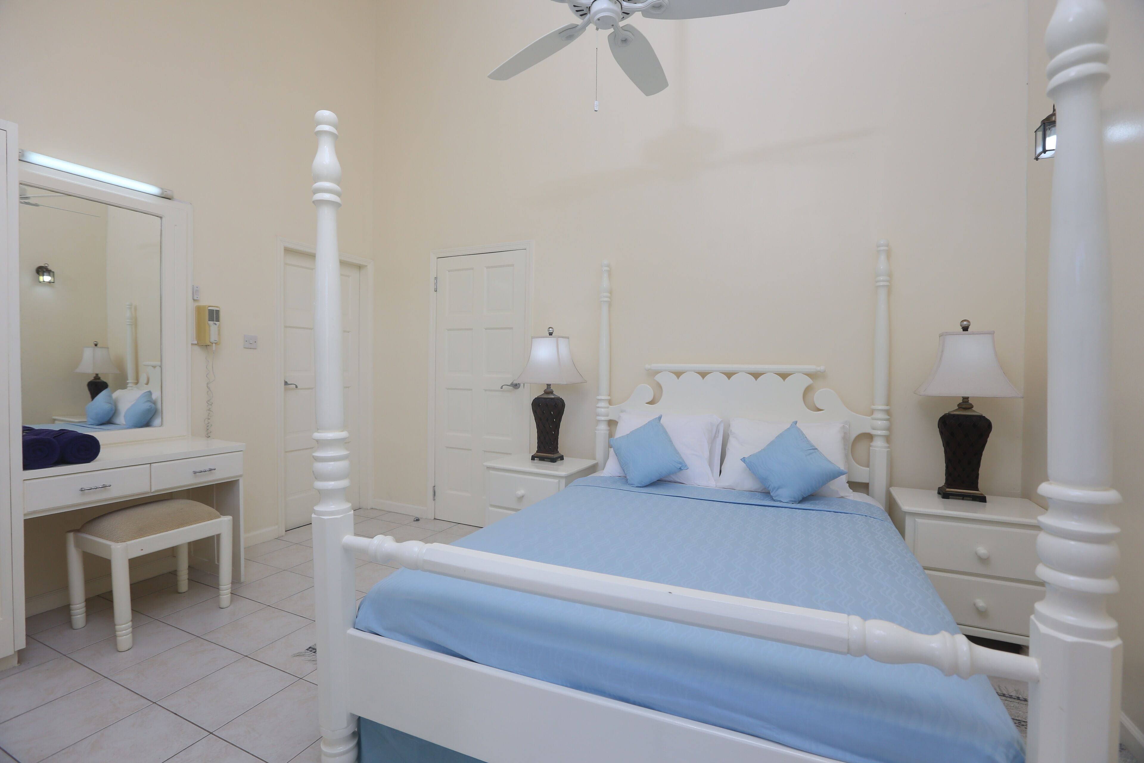A four-poster bed with a blue bedspread, a white dresser with a mirror, and two bedside tables with lamps.
