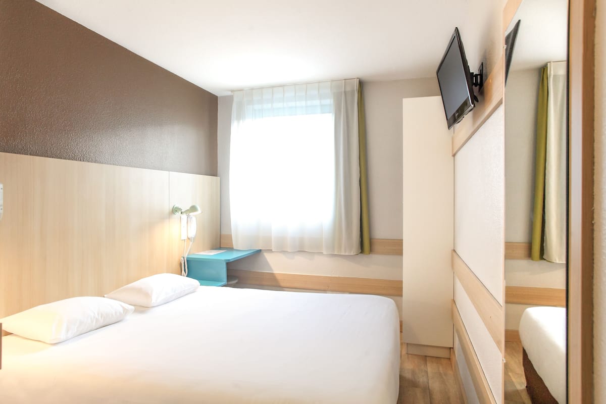 Una habitación de hotel con una cama, una mesita de noche, un televisor montado en la pared y una ventana con cortinas.