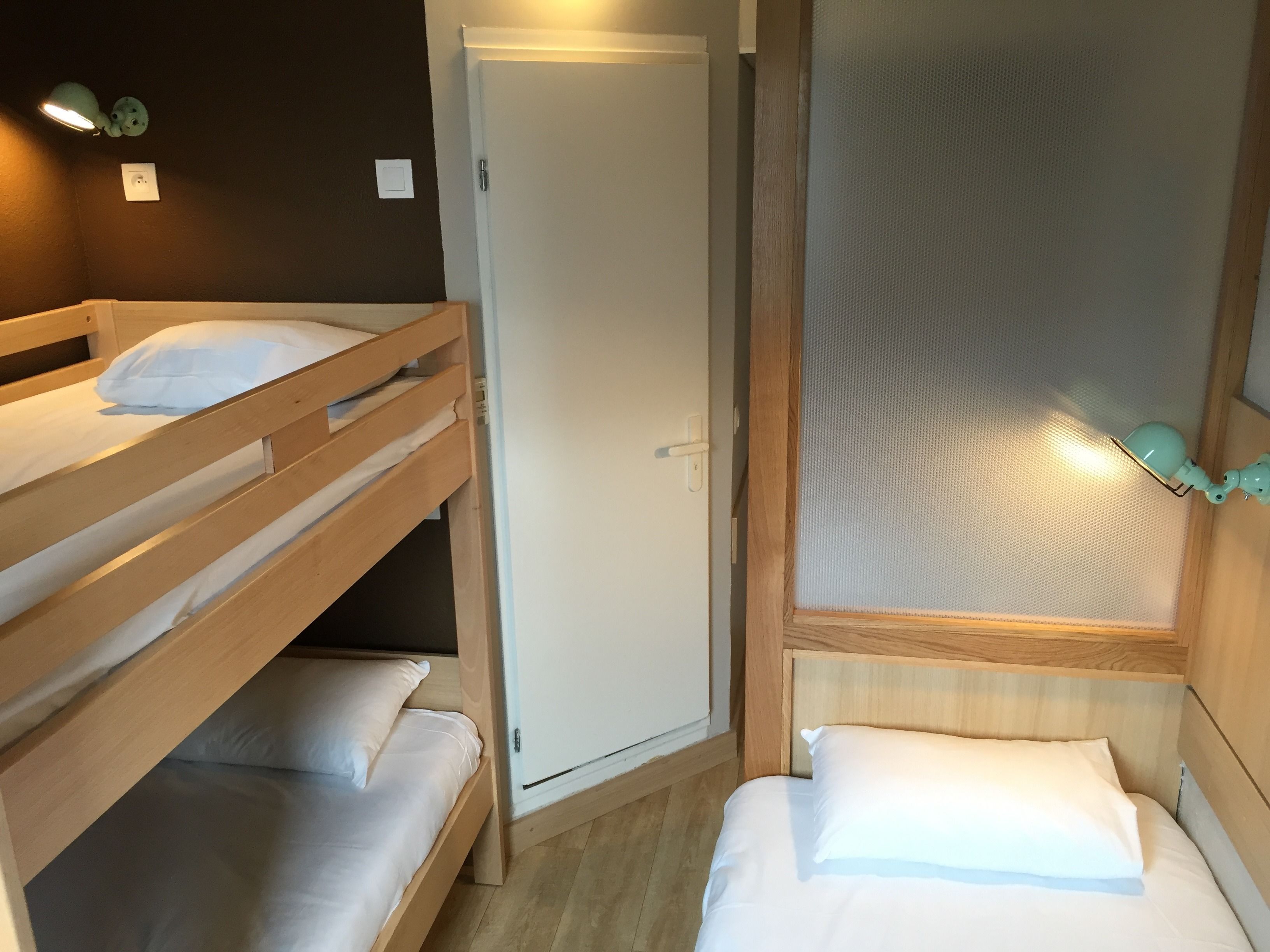 Quarto com beliche, porta, pequena janela com cortina e luminária fixada na parede.
