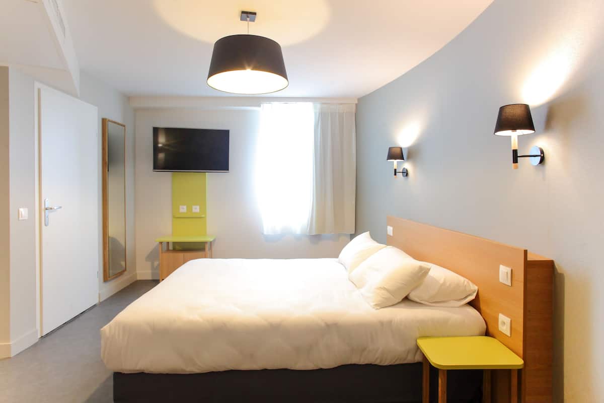 Una habitación de hotel moderna con una cama grande, mesitas de noche, un televisor de pantalla plana y un ventanal con cortinas.