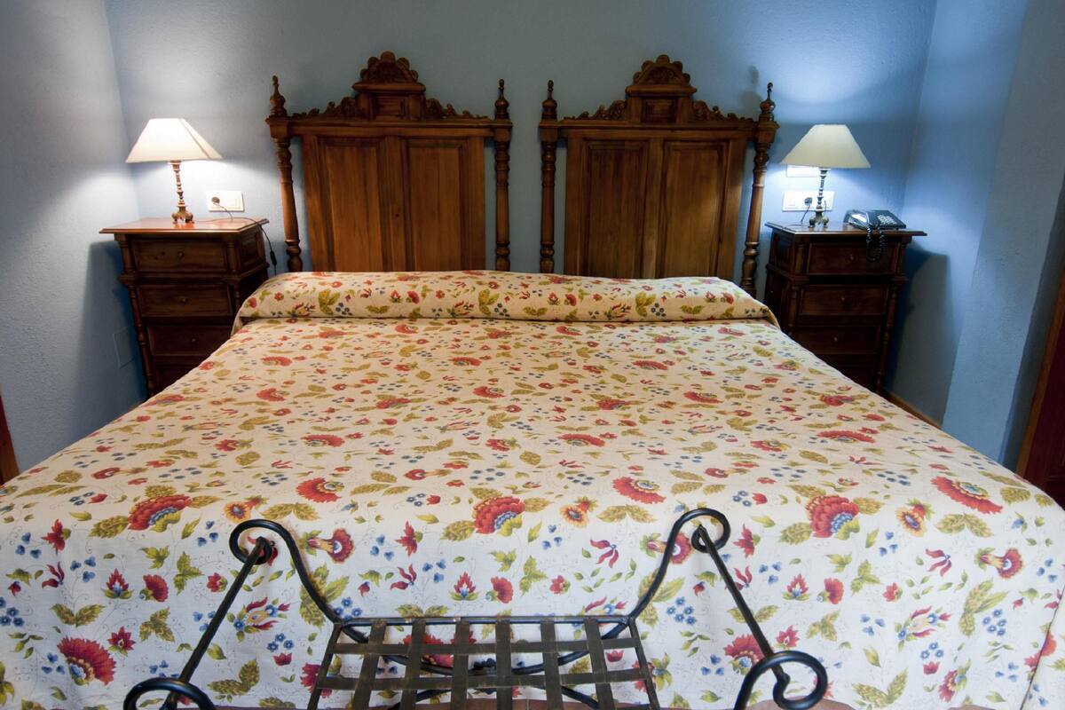 Un llit amb llençols florals, un capçal de fusta, dues taules de nit amb làmpades i una estructura de metall.