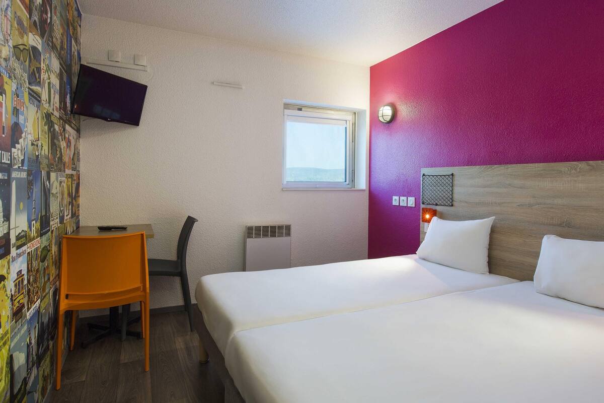Ein Hotelzimmer mit Bett, Schreibtisch, Sessel, Fernseher, Fenster und einer Wand mit einem Poster.