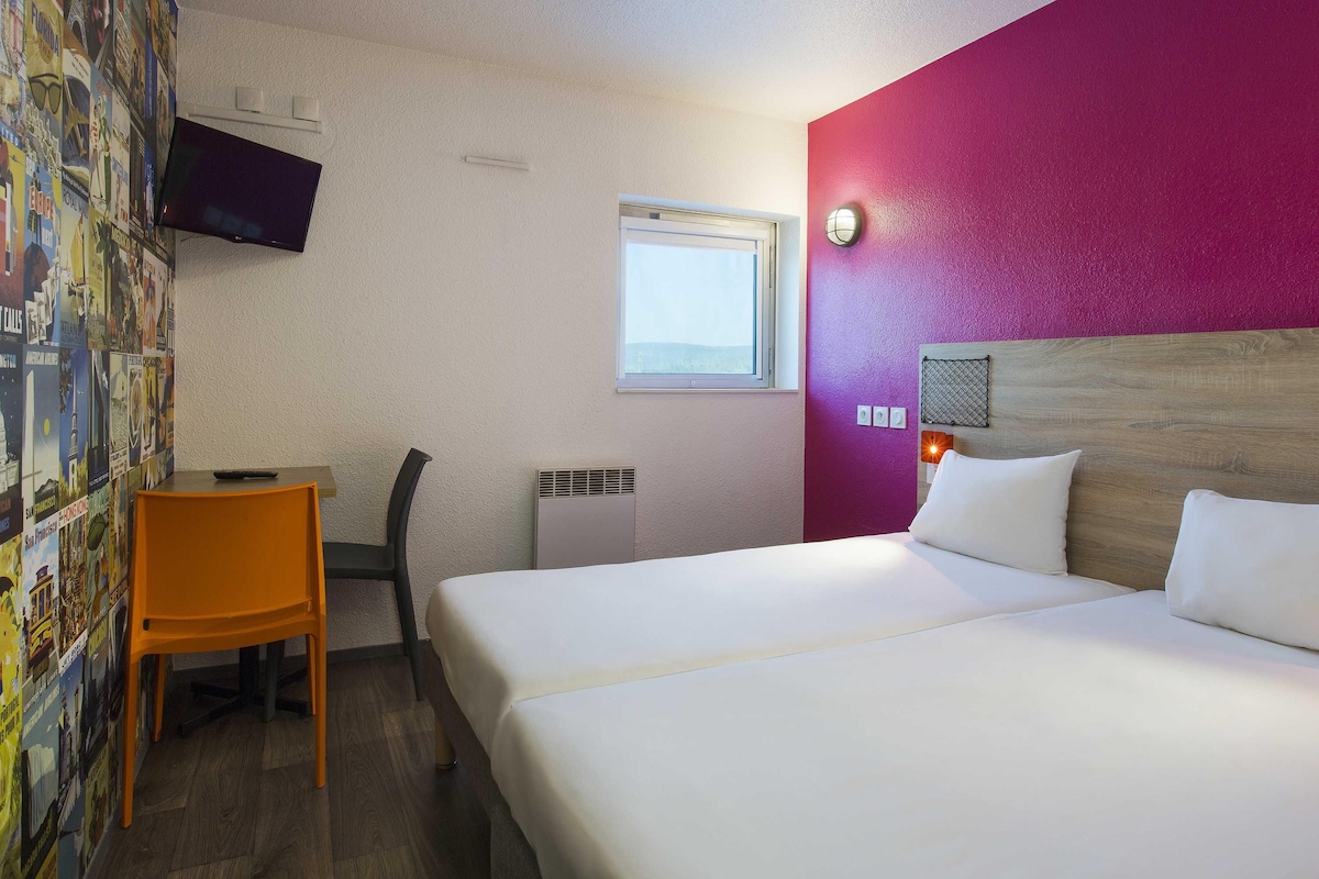Hotelkamer met een bed, een bureau, een stoel, een televisie, een raam en een muur met een poster.
