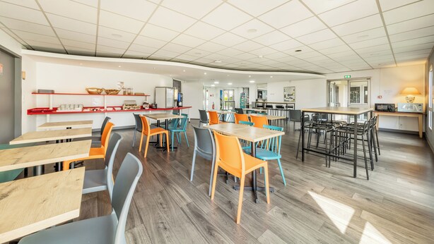 Un'ampia sala da pranzo con diversi tavoli e sedie, una cucina sullo sfondo e un design pulito e moderno.