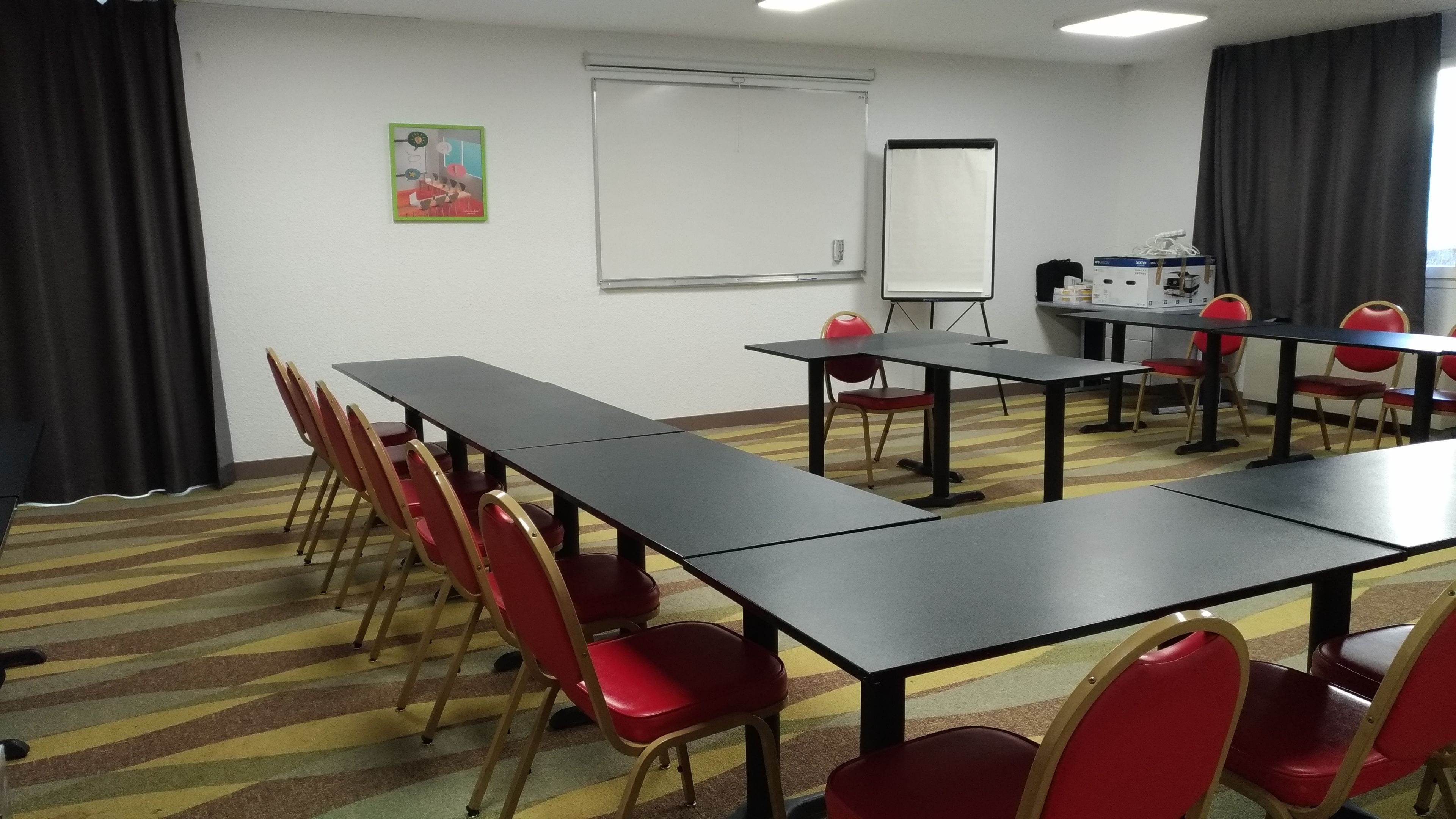 A conference room with long tables and red chairs, a whiteboard, and a projector screen.