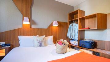 Quarto de hotel com cama, mesa de cabeceira, luminária, guarda-roupa e mala.