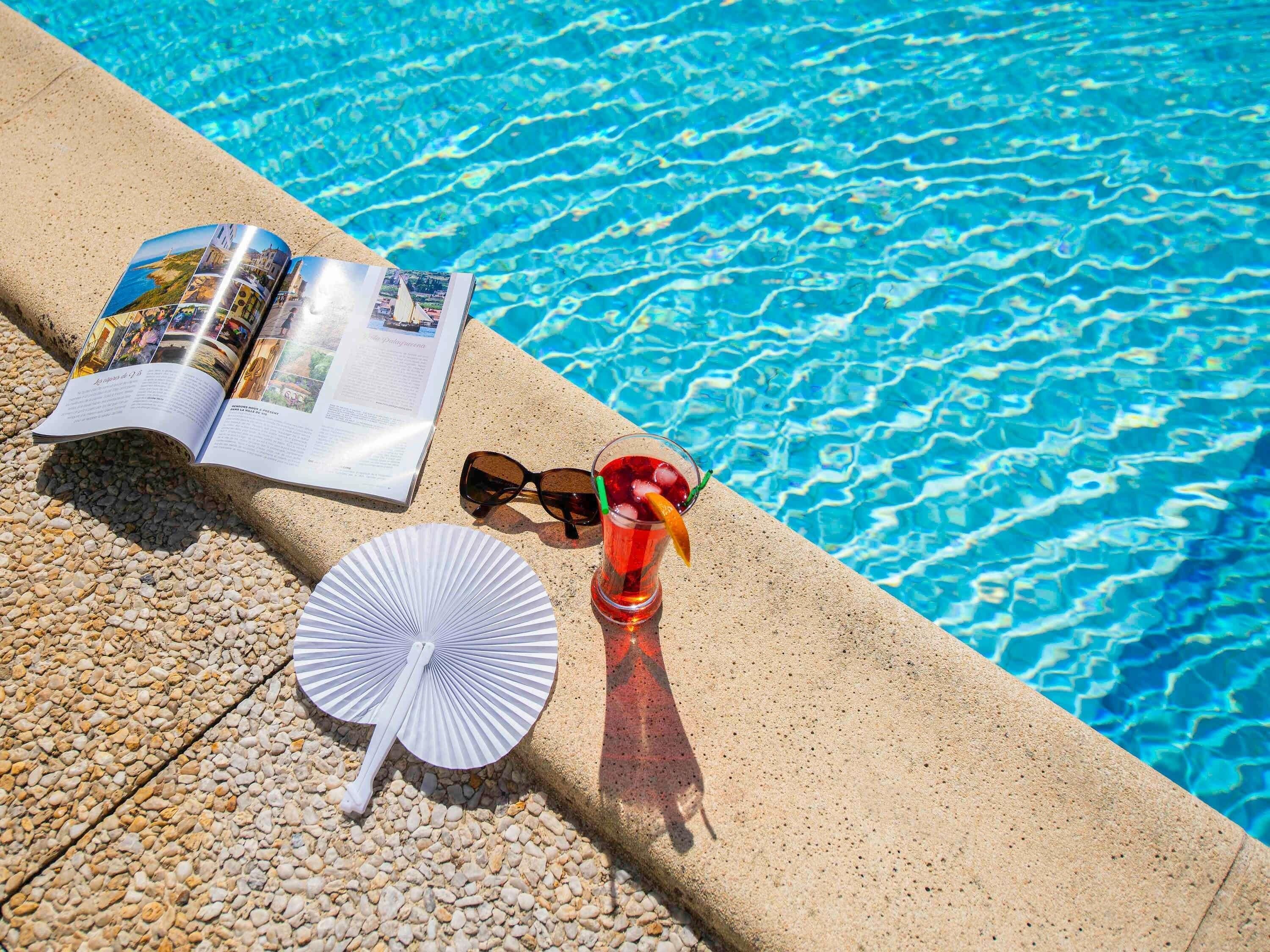 Escena junto a la piscina con un libro, gafas de sol, una bebida y un ventilador.
