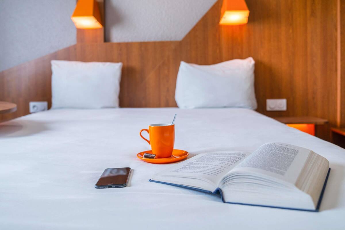 Une chambre d’hôtel avec un lit, un livre, une tasse et un téléphone intelligent posés sur le lit.