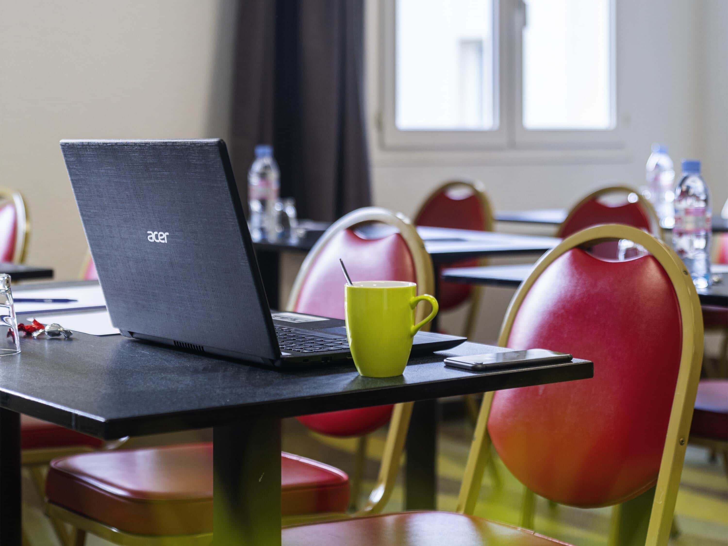 A laptop on a table with a yellow mug, red chairs, and a window in the background.