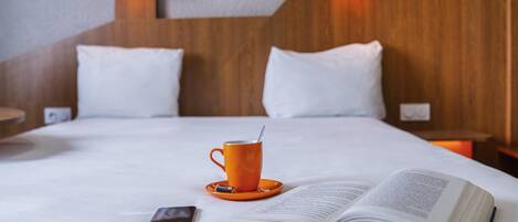 A neatly made bed with white linens, a book, a smartphone, and an orange cup on a white table.