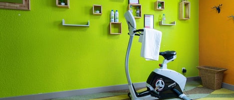 Una habitación con una bicicleta estática, un cesto de basura pequeño y varios objetos enmarcados en la pared.