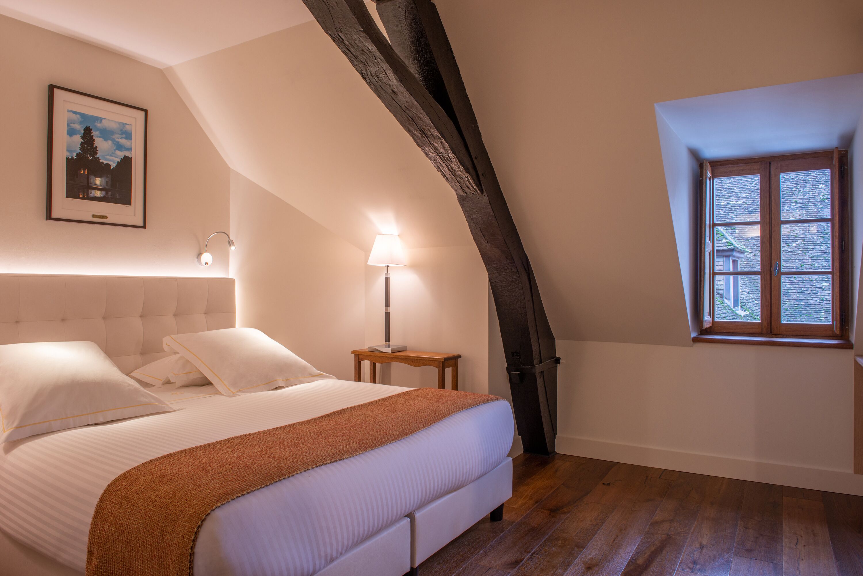 Un dormitorio con cama, mesita de noche, lámpara, un cuadro en la pared y ventana con vistas.