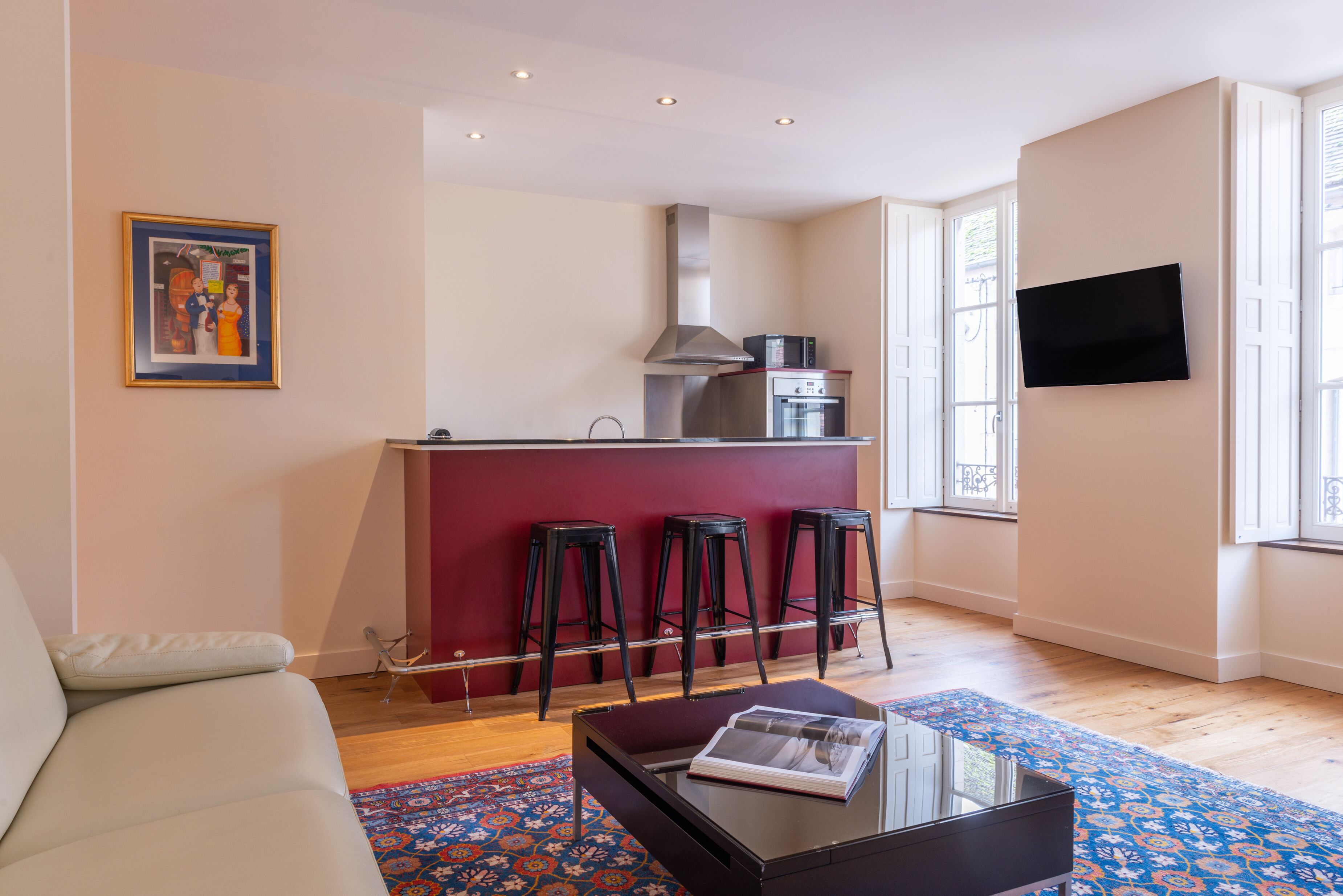 A modern living room with a sofa, a coffee table with a magazine, a red accent wall, a kitchen area with a stove and bar stools, and a TV mounted on the wall.