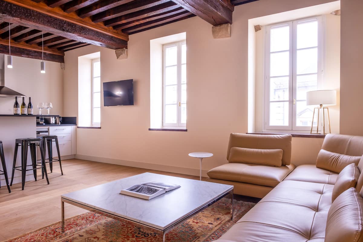 A modern living room with a sofa, a coffee table, and a TV. There are windows and a kitchen area visible.